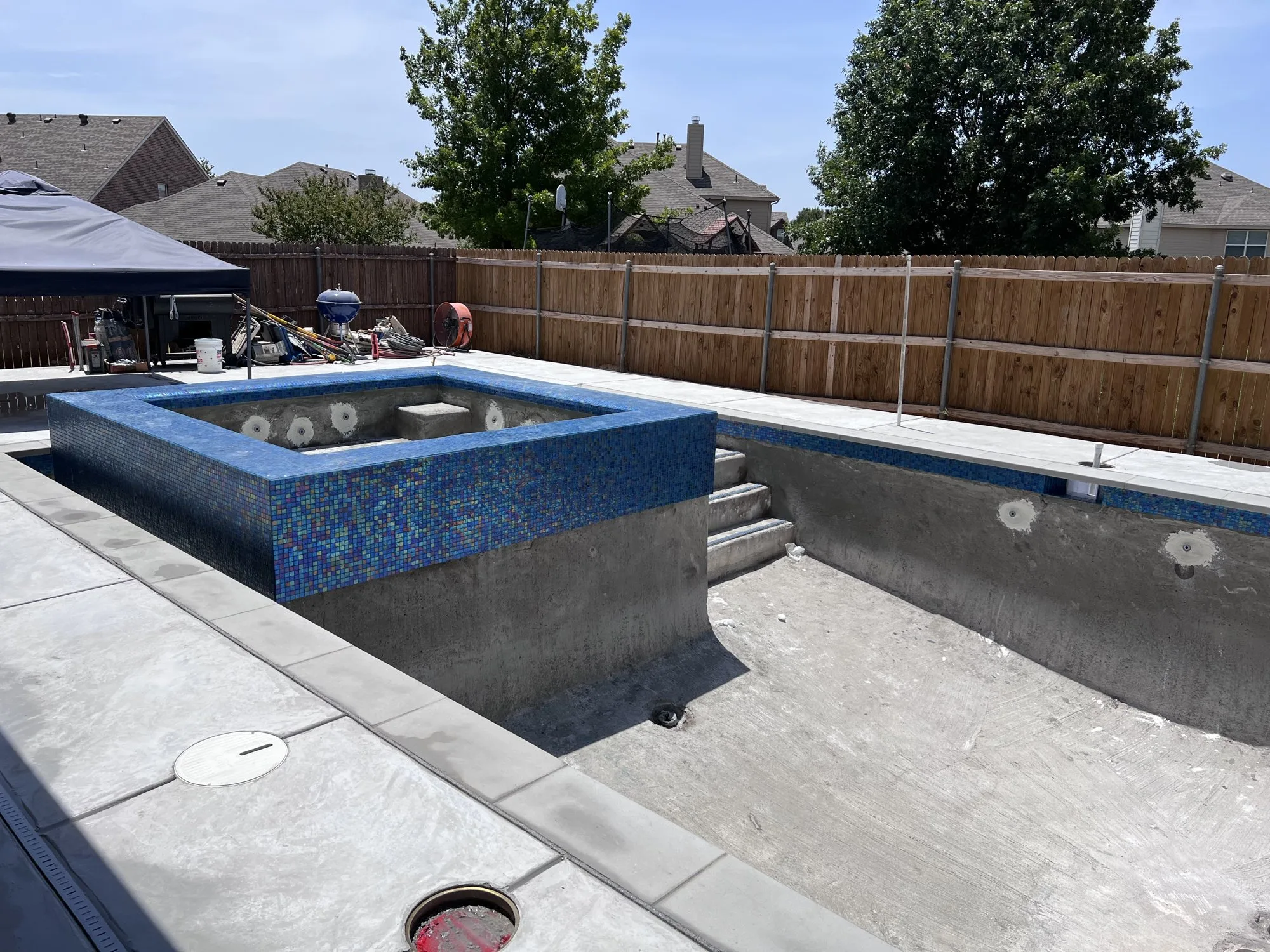 Raised spa with blue glass tile beside an empty pool shell during a Lucent pool build